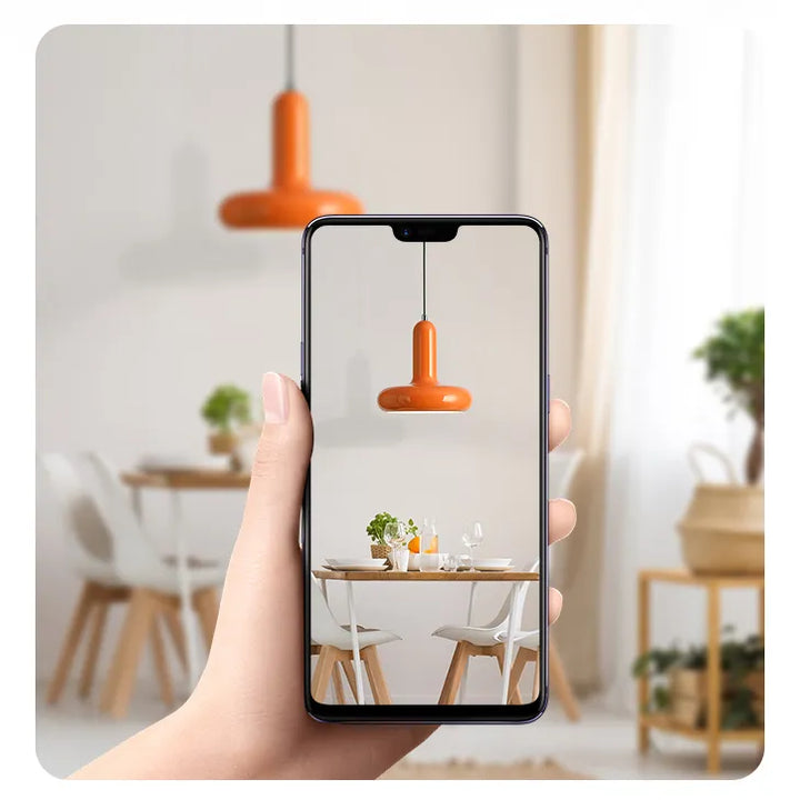 Une main tenant un téléphone avec une table et des chaises, une lampe sur la table, une plante floue, une chaise floue.