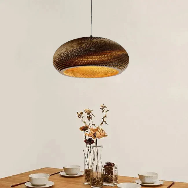 Une table avec un vase de fleurs et une suspension lumineuse en papier carton alvéolé globe aplati design scandinave, idéale pour une ambiance chaleureuse dans votre salon ou cuisine. Ampoule non incluse. Dimensions : 40x17 cm.